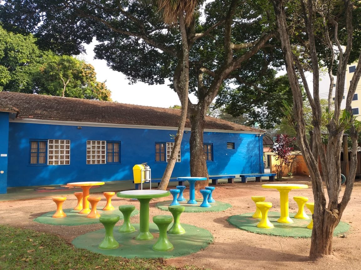 Mesas e bancos coloridos sob as árvores no espaço de convivência da Escola Coopep